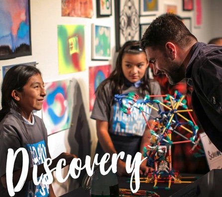 Two young girls in dark gray T-Shirts work on an intricate model which an instructor looks on, smiling. Colorful artwork adorns the walls behind them.