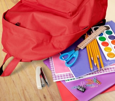 Red backpack on a table beside a stapler, notebooks, folders, paints, pencils, scissors and a ruler