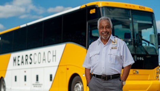 Smiling Motorcoach Operator stands beside a yellow and white Mears coach bus.