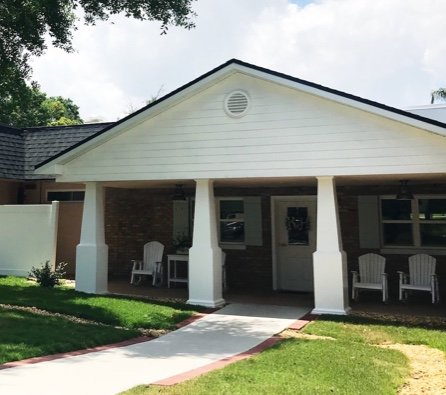 Wide front porch with white columns on a one-story building holds three white chairs on either side of the front door