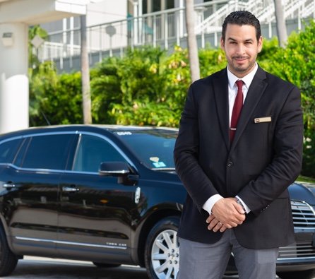 Handsome, smiling luxury private car service chauffeur in black jacket, white shirt, maroon tie and grey pants stands beside a black Mears luxury sedan